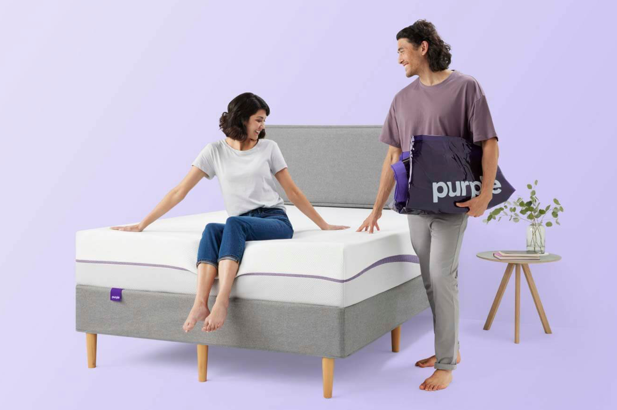 Woman sitting on Purple Plus Mattress while man standing next to it