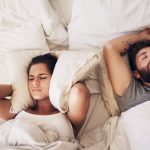 A young woman covering her ears with a pillow while her husband snores in bed