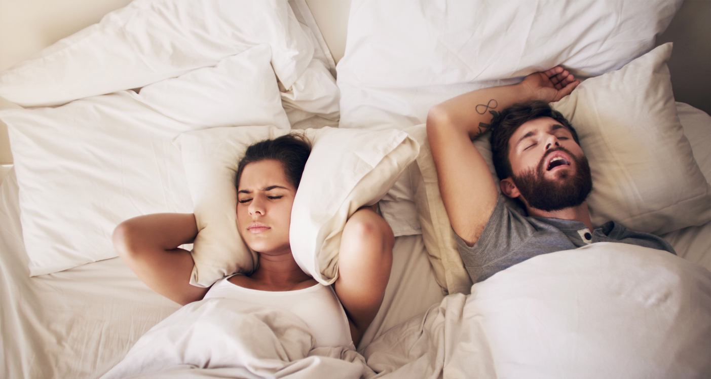 A young woman covering her ears with a pillow while her husband snores in bed